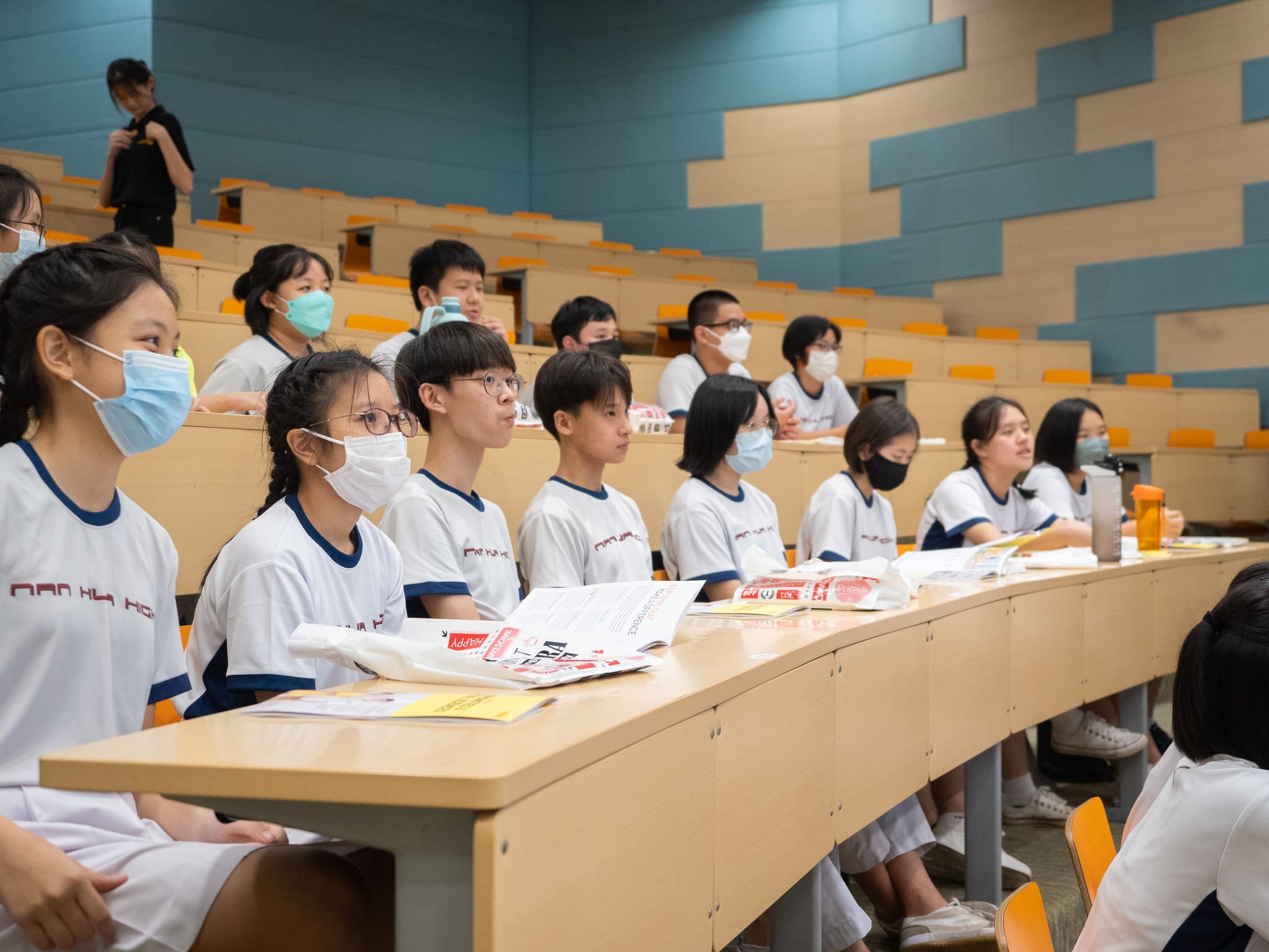 Students in white uniforms and face masks sit attentively in tiered lecture hall seating with papers on desks.